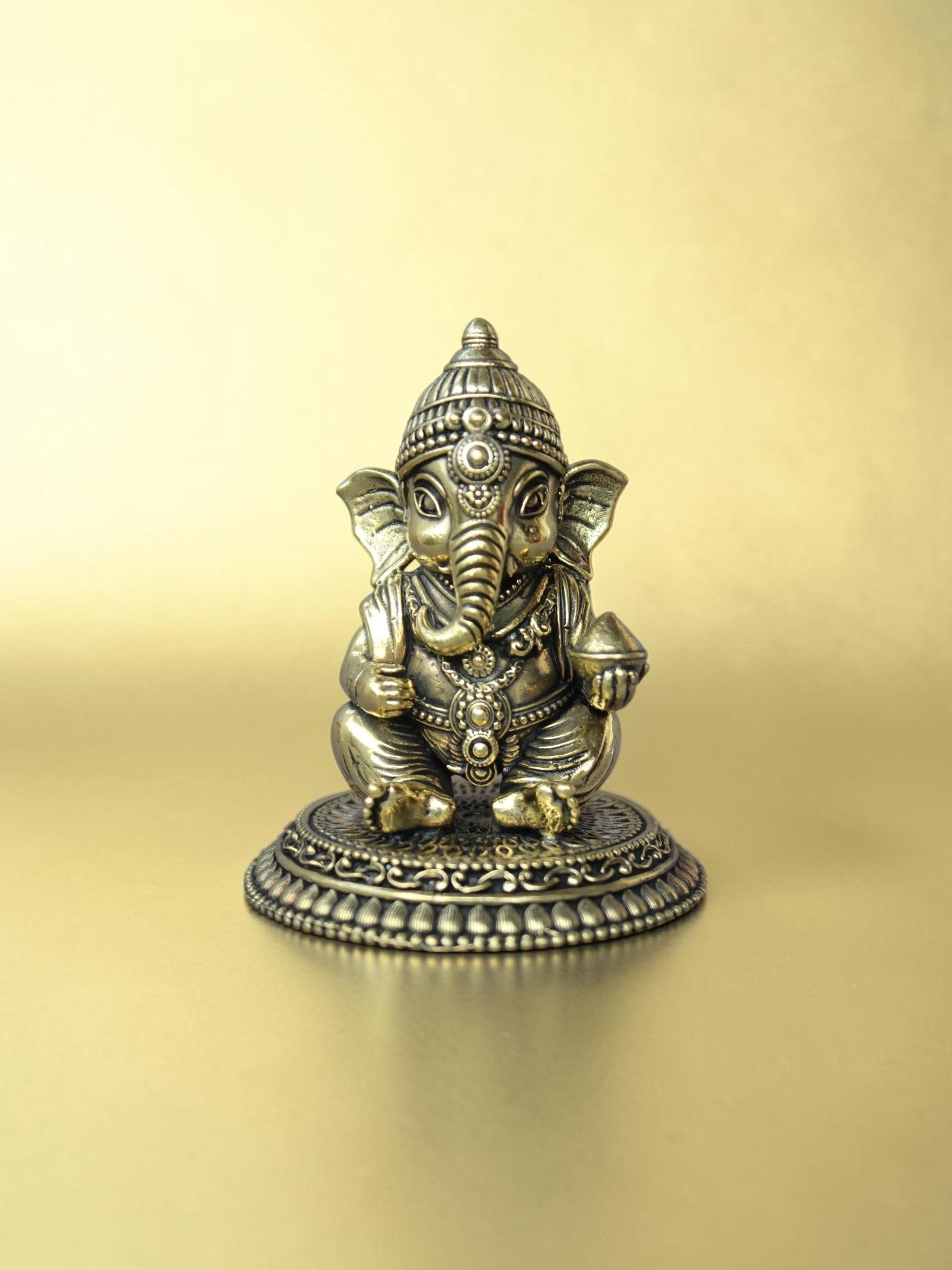 Rigvedaa™ handcrafted brass Bal Ganesha idol in a seated pose with an antique gold finish on an ornate pedestal.