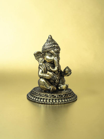 Rigvedaa™ handcrafted brass Bal Ganesha idol in a seated pose with an antique gold finish on an ornate pedestal.