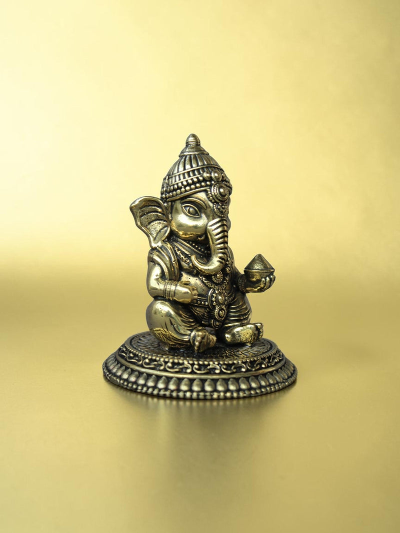 Rigvedaa™ handcrafted brass Bal Ganesha idol in a seated pose with an antique gold finish on an ornate pedestal.
