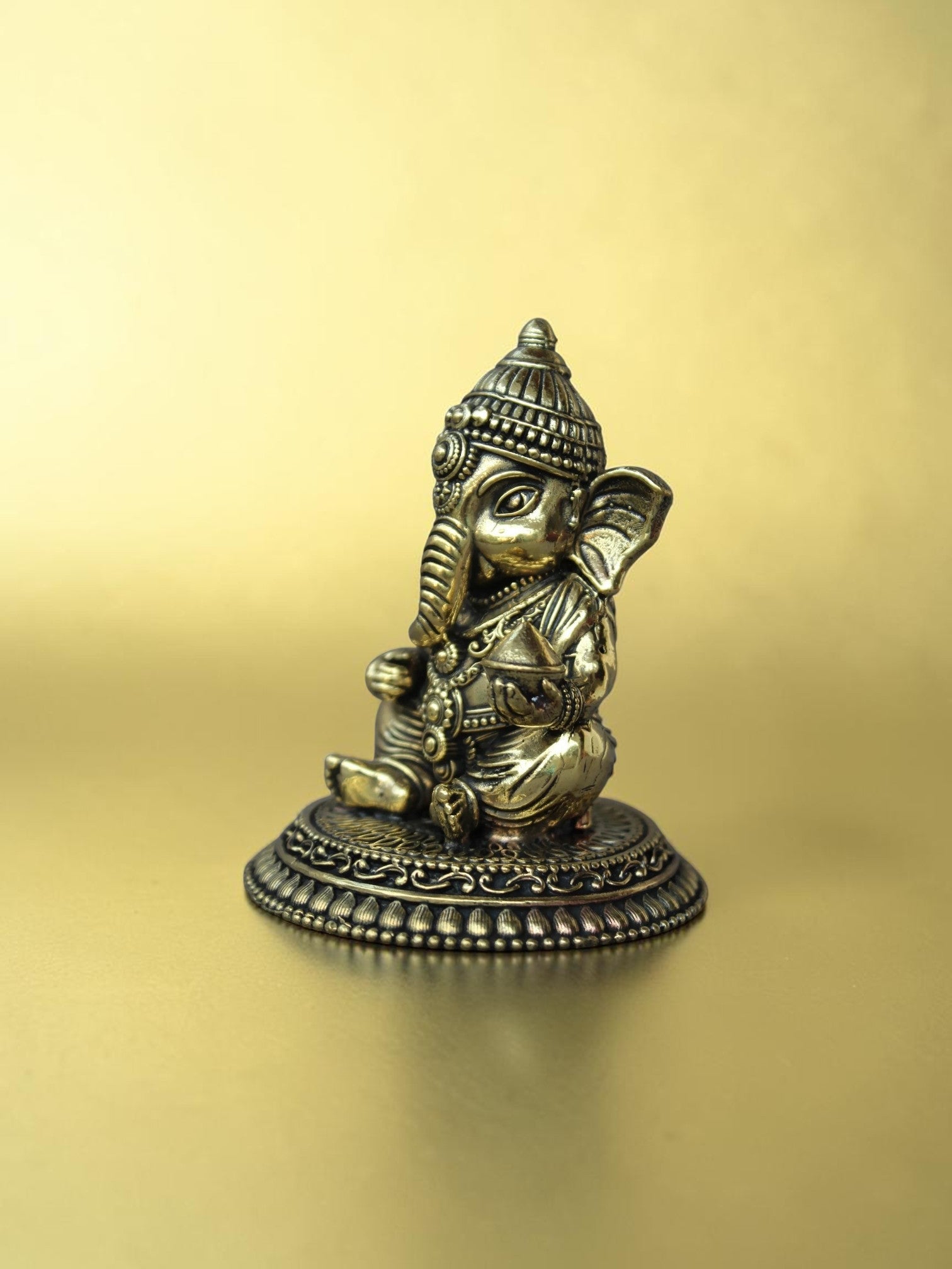 Rigvedaa™ brass Bal Ganesha idol in a seated pose with an antique gold finish on an ornate circular pedestal.
