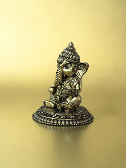 Rigvedaa™ brass Bal Ganesha idol in a seated pose with an antique gold finish on an ornate circular pedestal.