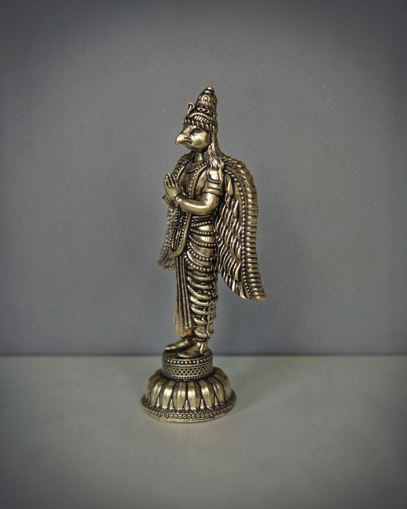 Rigvedaa brass Garuda statue with folded hands, detailed wings, and ornate base. Antique gold finish.