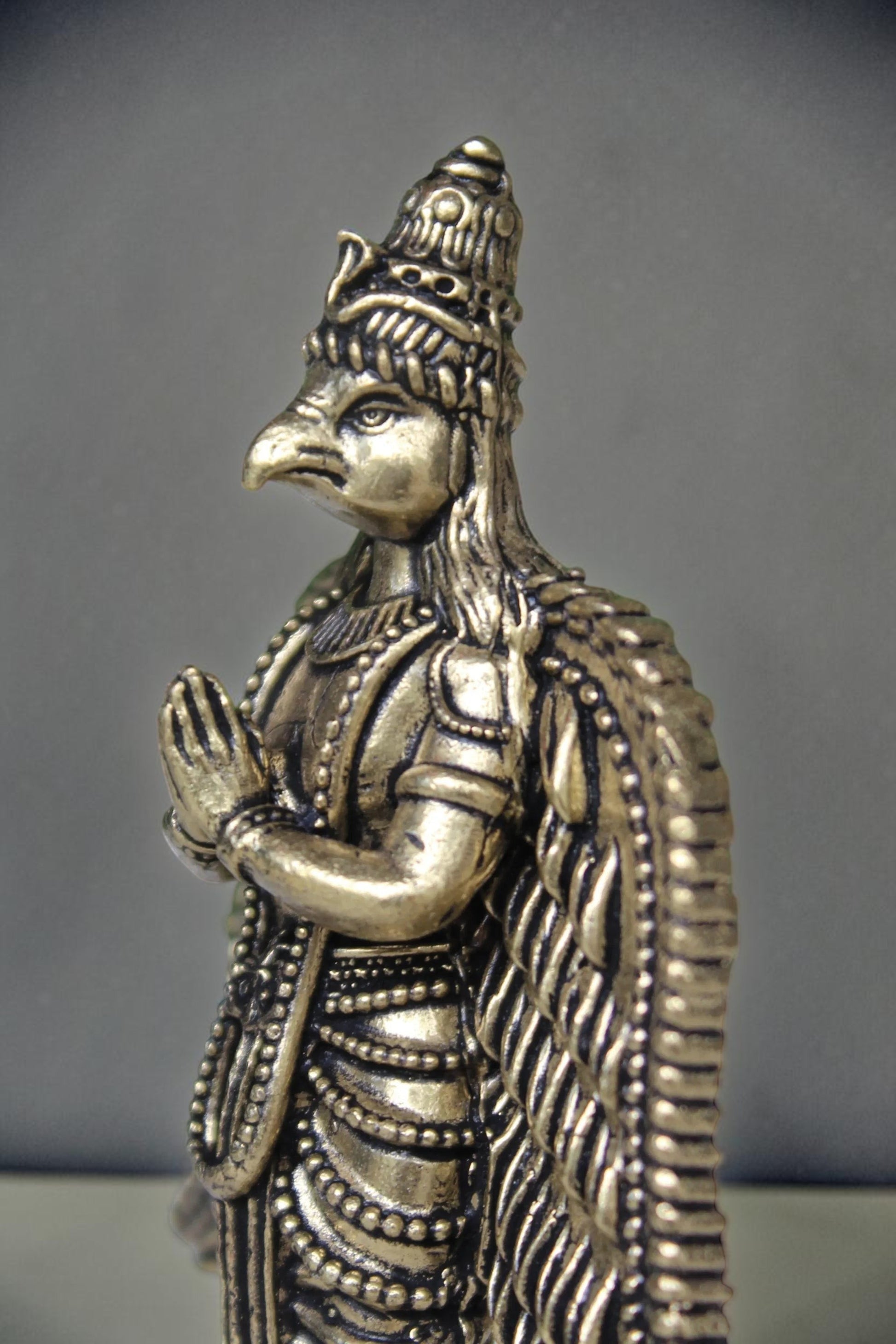 Rigvedaa brass Garuda statue with folded hands, detailed wings, and ornate headdress. Antique gold finish.