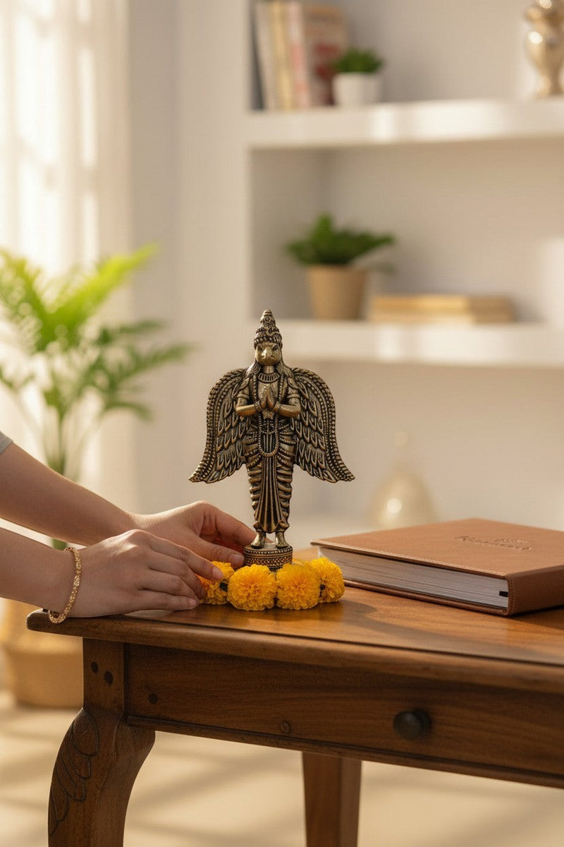 Rigvedaa brass Garuda statue with hands in prayer pose, spread wings, and intricate details. Yellow marigolds adorn the base.