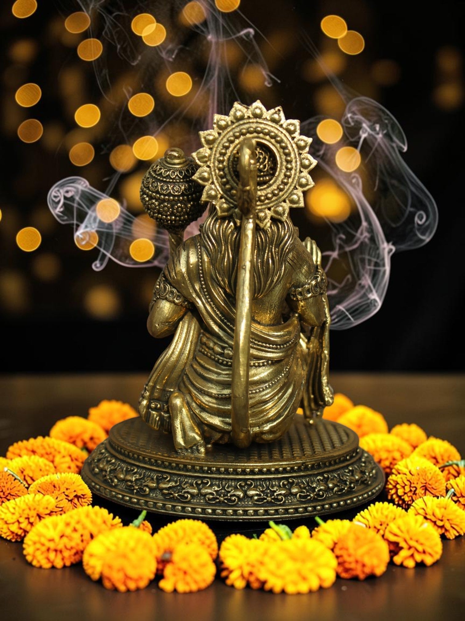 Rigvedaa™ brass Lord Hanuman idol seated on a pedestal with a mace, surrounded by marigold flowers and incense smoke.