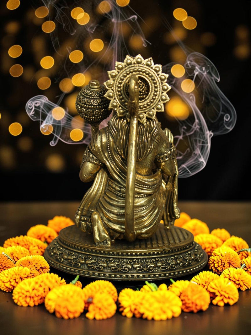 Rigvedaa™ brass Lord Hanuman idol seated on a pedestal with a mace, surrounded by marigold flowers and incense smoke.