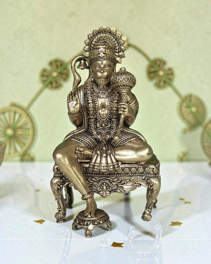 Rigvedaa™ brass Hanuman Ji idol sitting on an ornate throne, holding a mace and bell. Features intricate detailing and a golden finish.