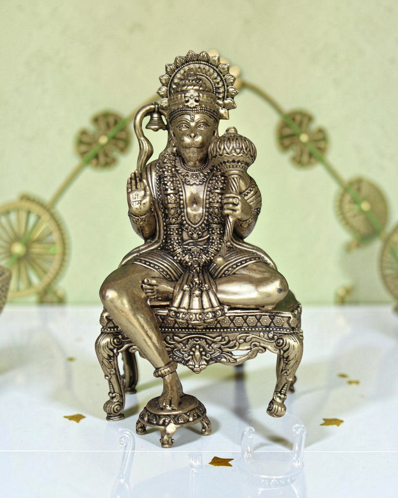 Rigvedaa™ brass Hanuman Ji idol sitting on an ornate throne, holding a mace and bell. Features intricate detailing and a golden finish.