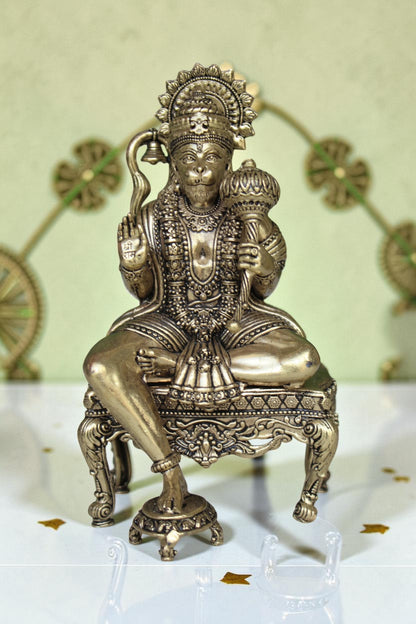 Rigvedaa™ brass Hanuman Ji idol sitting on an ornate throne, holding a mace and bell. Features intricate detailing and a golden finish.