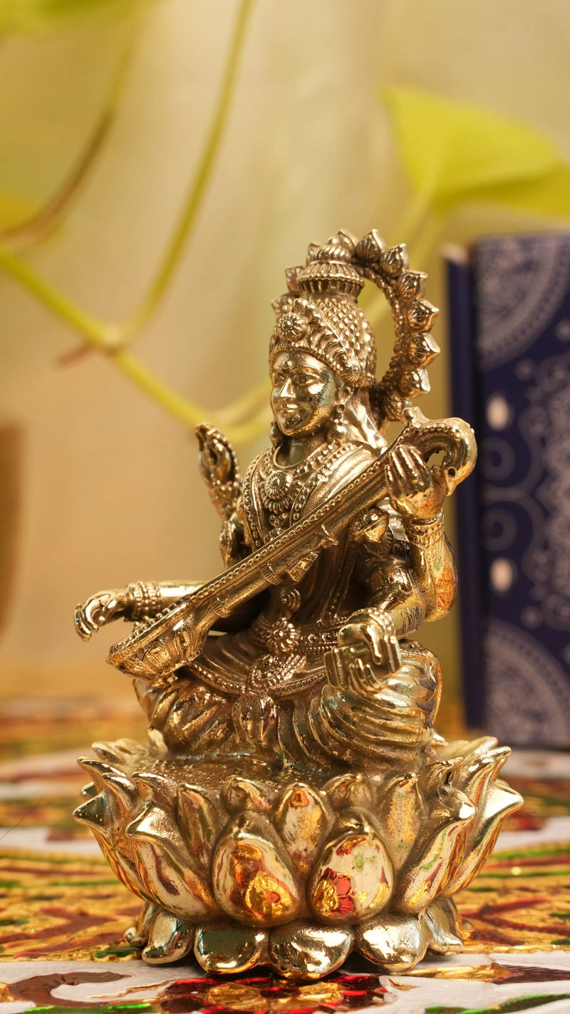 Rigvedaa™ brass Saraswati idol seated on lotus, holding veena and scriptures. Detailed, golden finish, divine sculpture.