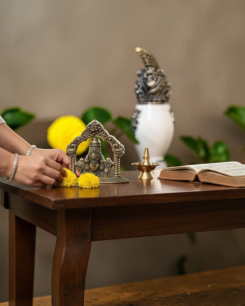 Rigvedaa™ brass Lord Venkateswara Tirupati Balaji face idol with intricate crown on a wooden table with marigold flowers.