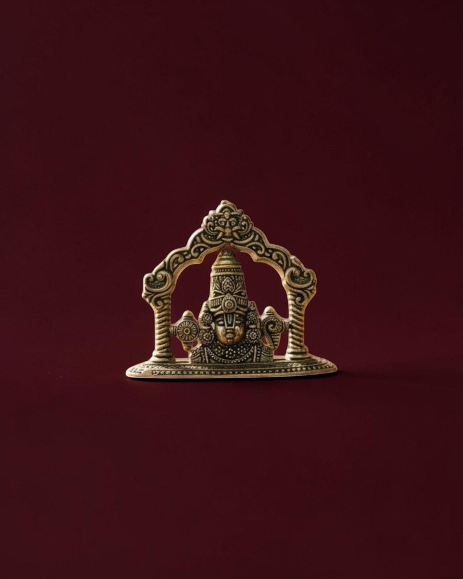 Rigvedaa™ brass Lord Venkateswara Tirupati Balaji face idol with intricate crown detailing on a dark red background.
