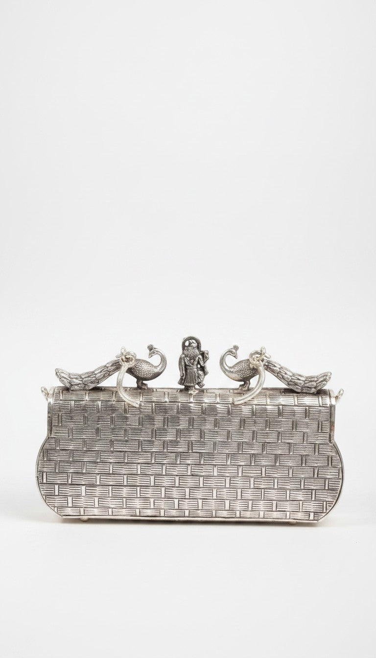 Rigvedaa™ Leela silver plated brass clutch with intricate carvings, peacock accents, and a central figurine. Woven texture.