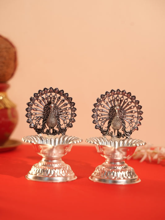 Two Rigvedaa™ silver-plated brass peacock diya oil lamps with intricate lotus bowls on a red surface for pooja rituals.