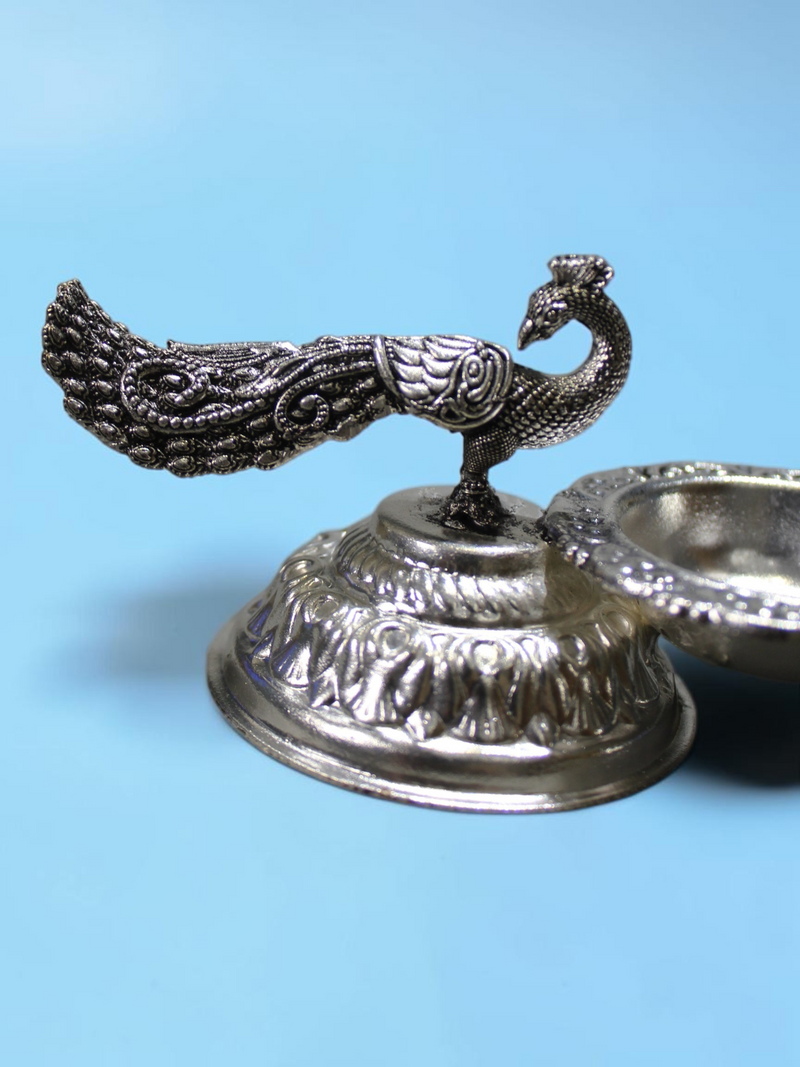 Rigvedaa™ silver plated brass peacock diya oil lamp with intricate tail detailing on a lotus pedestal base.
