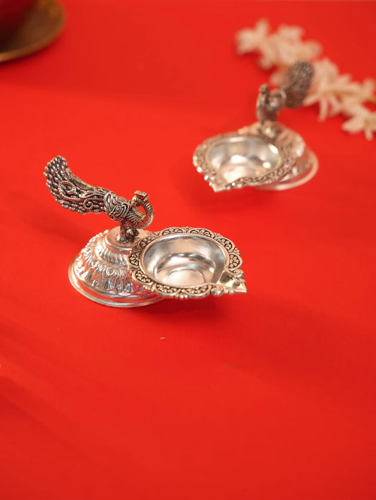 Rigvedaa™ silver plated brass peacock diya oil lamps with intricate tail detailing on a vibrant red background