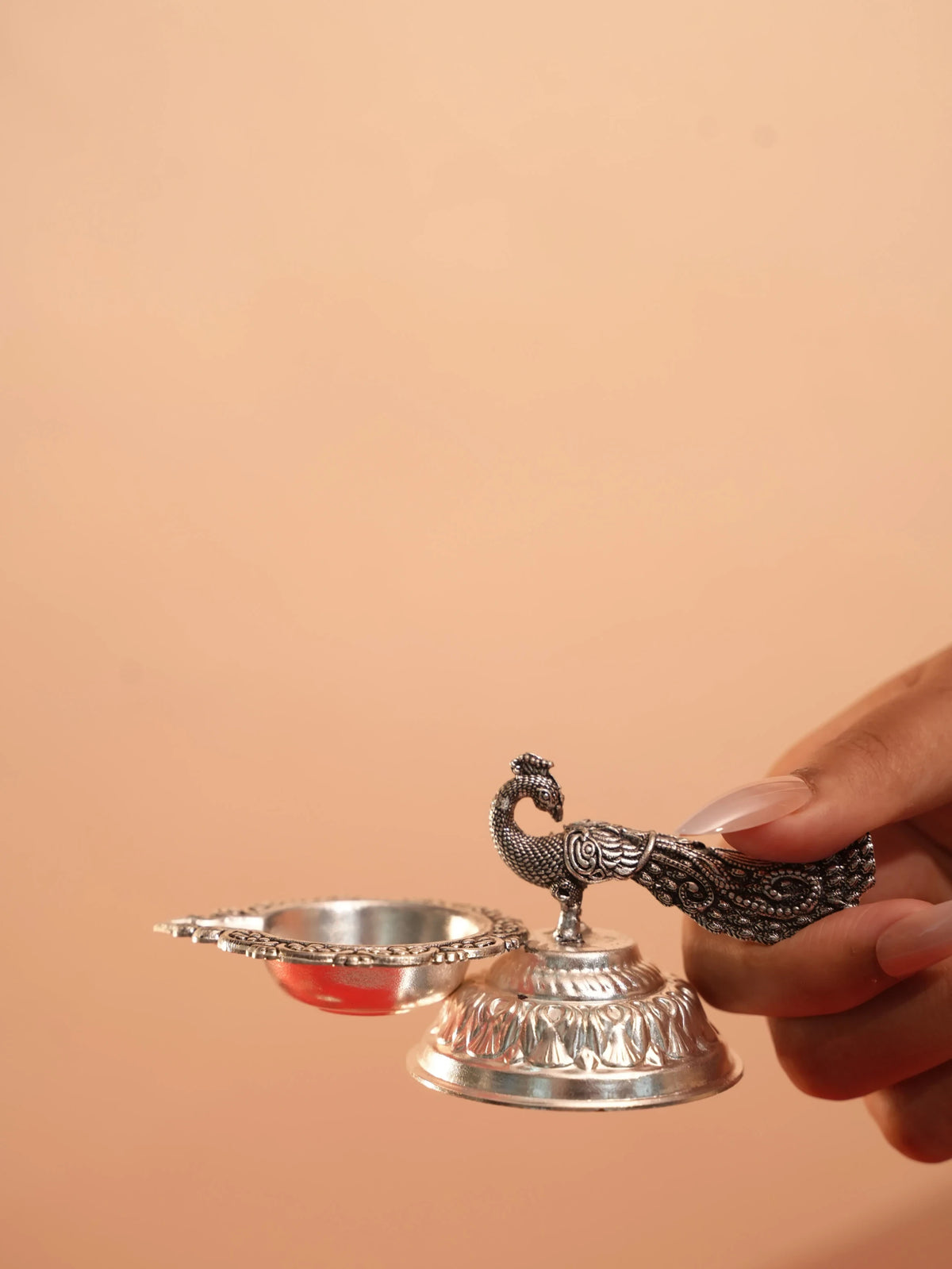 Rigvedaa™ silver plated brass peacock diya oil lamp held in hand, featuring intricate tail detailing and lotus base.