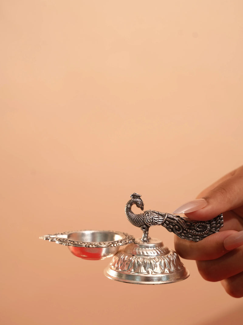 Rigvedaa™ silver plated brass peacock diya oil lamp held in hand, featuring intricate tail detailing and lotus base.