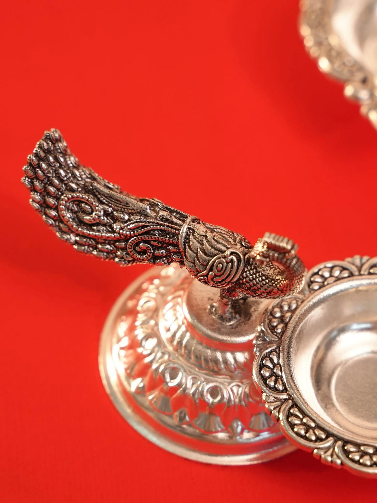 Rigvedaa™ silver plated brass diya featuring an intricate peacock tail design on a lotus pedestal against red fabric