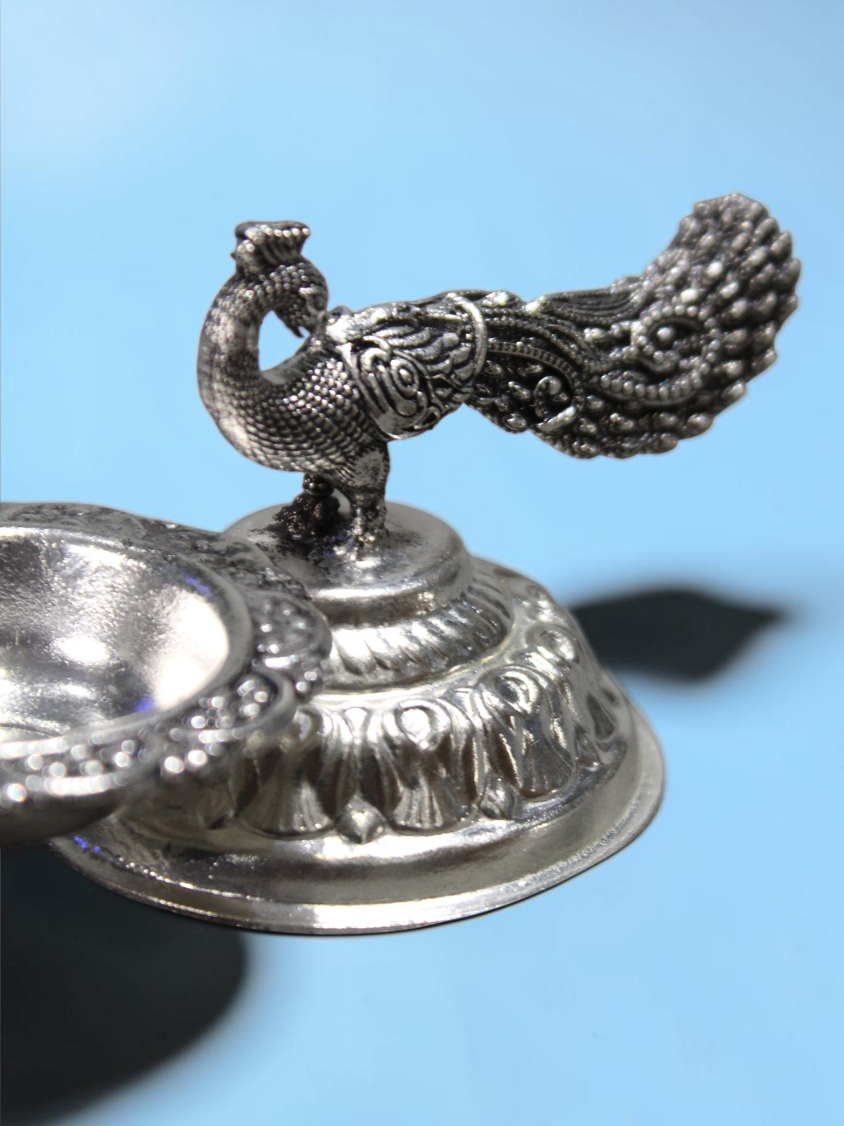 Rigvedaa™ silver plated brass diya featuring an intricate peacock design with a long tail and lotus pedestal base.